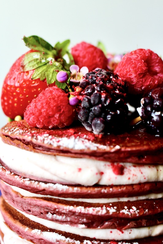 Pink pancakes stacked and filled with ricotta, covered with raspberries and blueberries and strawberries. Sprinkled with powder sugar and maple syrup.